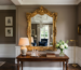 Ornate gold-framed mirror reflects chandelier and window over wooden console table with lamp and flower vase